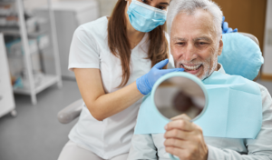 Elderly man smiling and holding a mirror up to his all-on-4 dental implants at Virginia Surgical Arts in Virginia Beach, VA