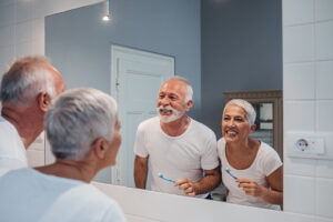 Older couple smiling and brushing their teeth in large mirror at Virginia Surgical Arts in Virginia Beach, VA